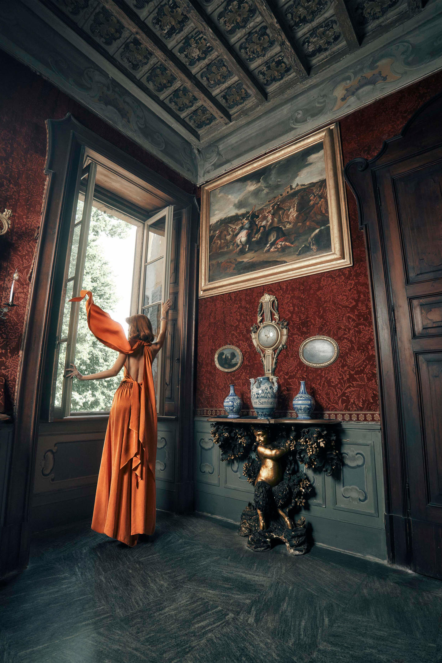 Woman in an orange outfit standing in a ornate room with red walls and decorative ceiling.