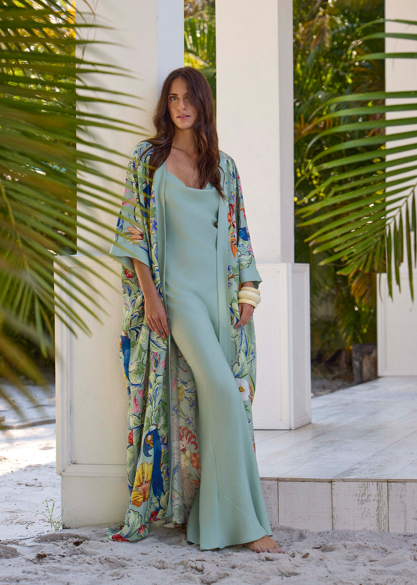 Model wearing a light green maxi dress with a printed kimono cover-up, standing barefoot outdoors near white columns and palm leaves.