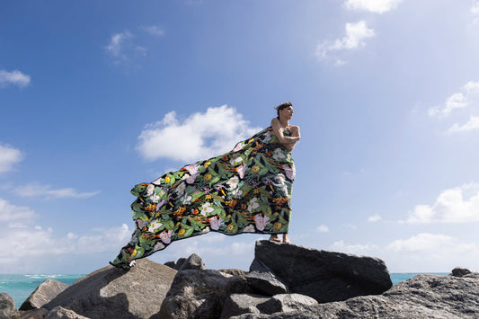 A model in a floral fabric piece from niLuu on ocean rocks, the fabric billowing in the wind against blue sky.