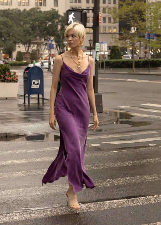 A woman wearing a violet slip dress with a cowl neck from niLuu, with short blonde hair, is walking down the road.