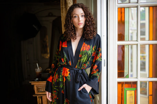 A woman with curly hair is wearing niLuu’s Olivia print vegan silk kimono robe and leaning against a wall.