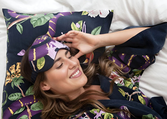 A woman smiles while sleeping in a niLuu purple snake print Jagger sleep mask and matching robe and pillowcase.
