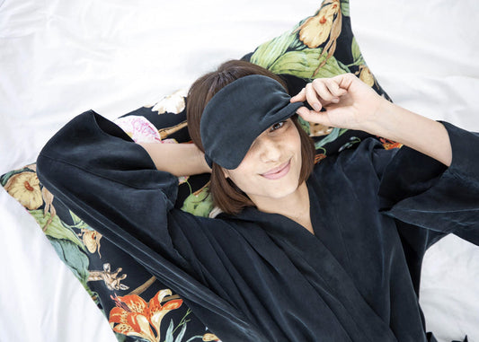 A woman wearing a black vegan silk robe and sleep mask, lying on a beautiful silk pillowcase, all from niLuu.