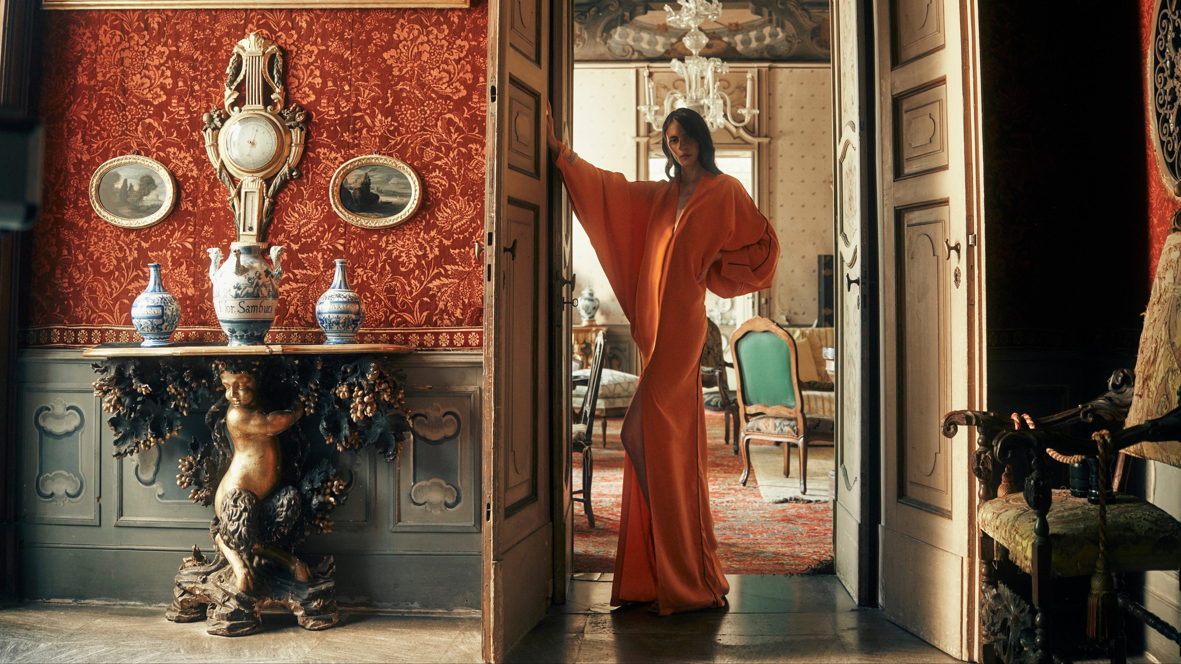 A woman wearing a flowing orange dress stands in an ornate interior doorway, with a decorative red patterned wall, antique-style console table, paintings, and furniture visible around her.