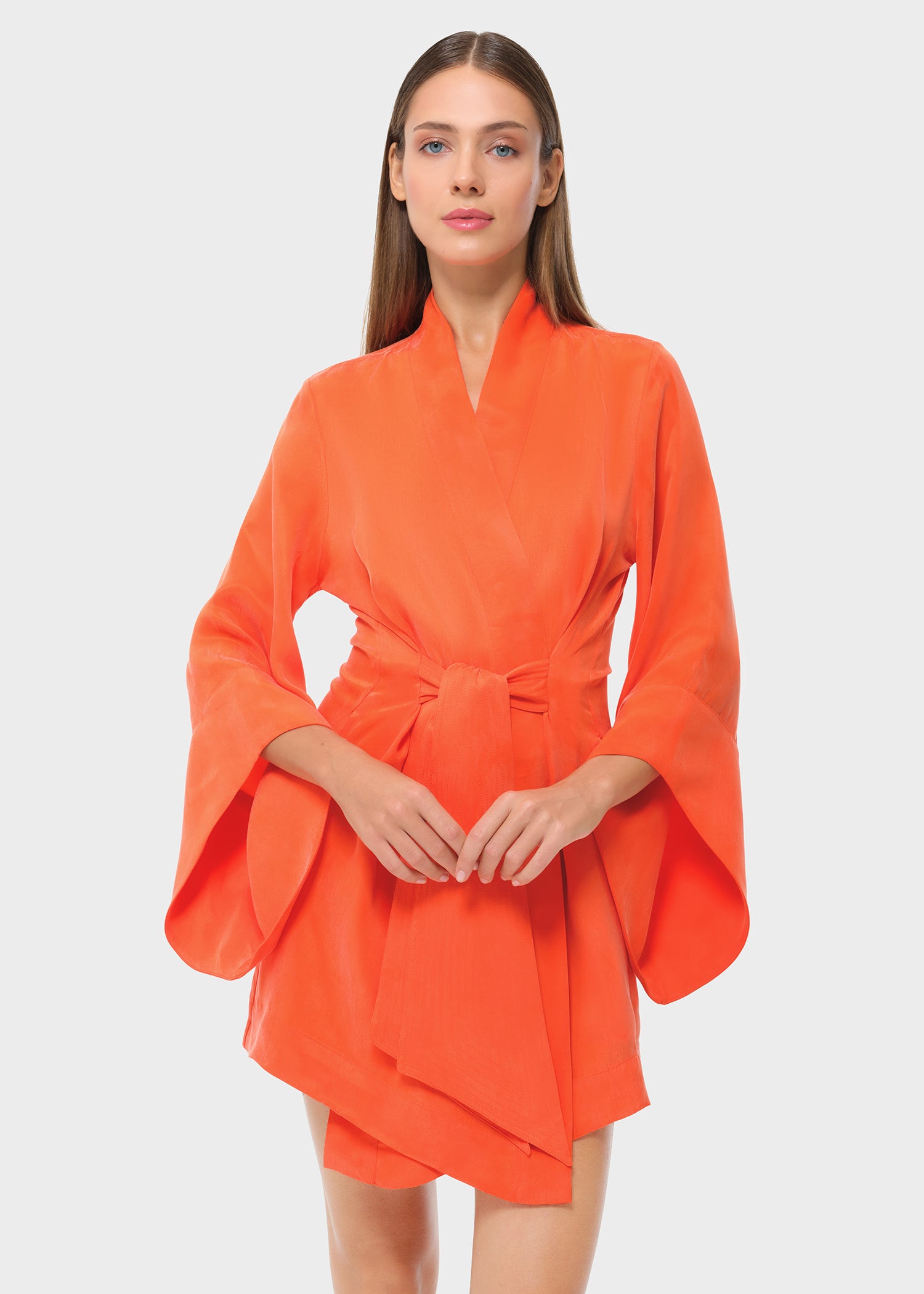 Front view of model wearing orange wrap mini dress with tie waist and wide sleeves, studio background