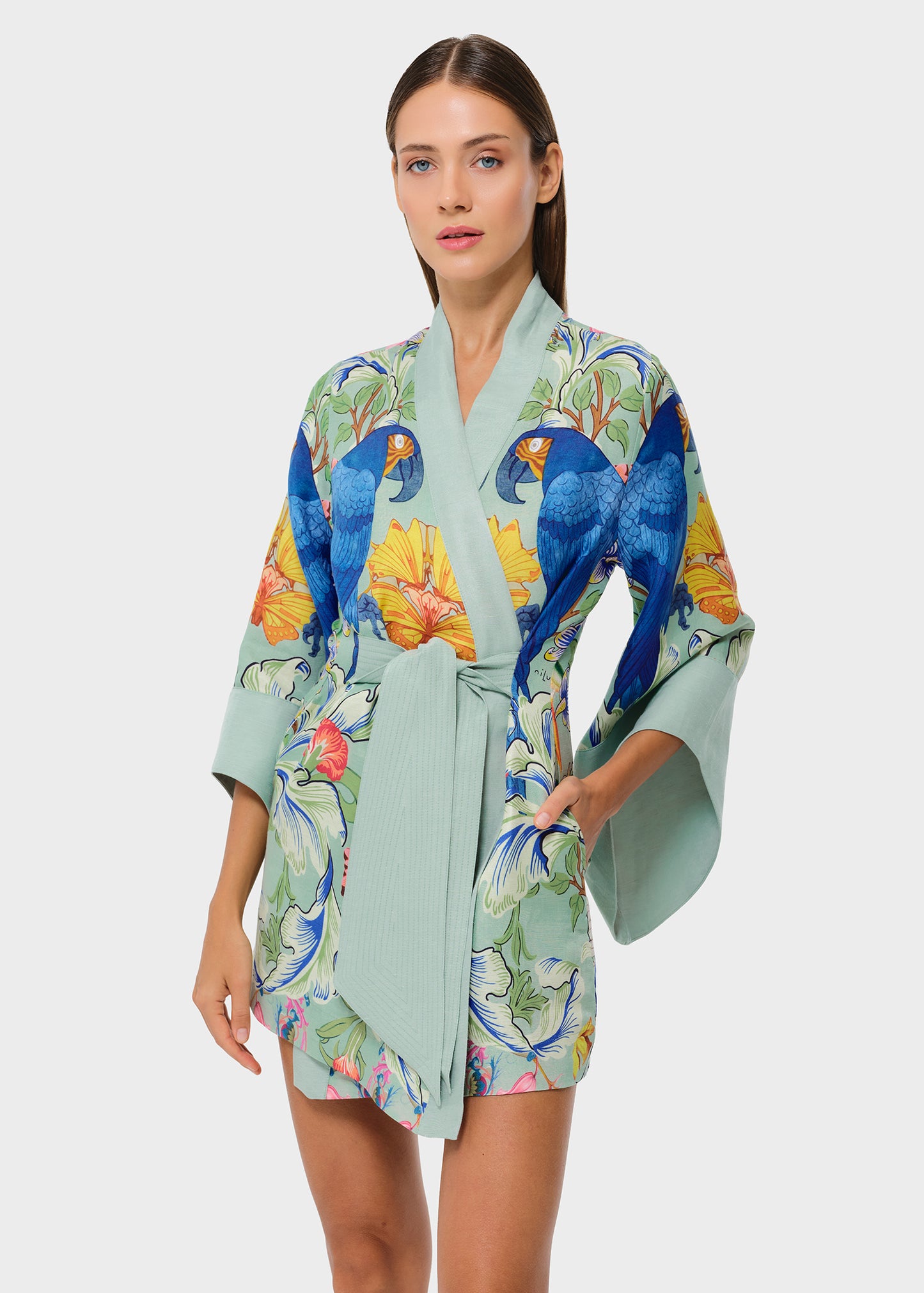Front view of model wearing a light green kimono-style wrap mini dress with blue parrot and floral print.