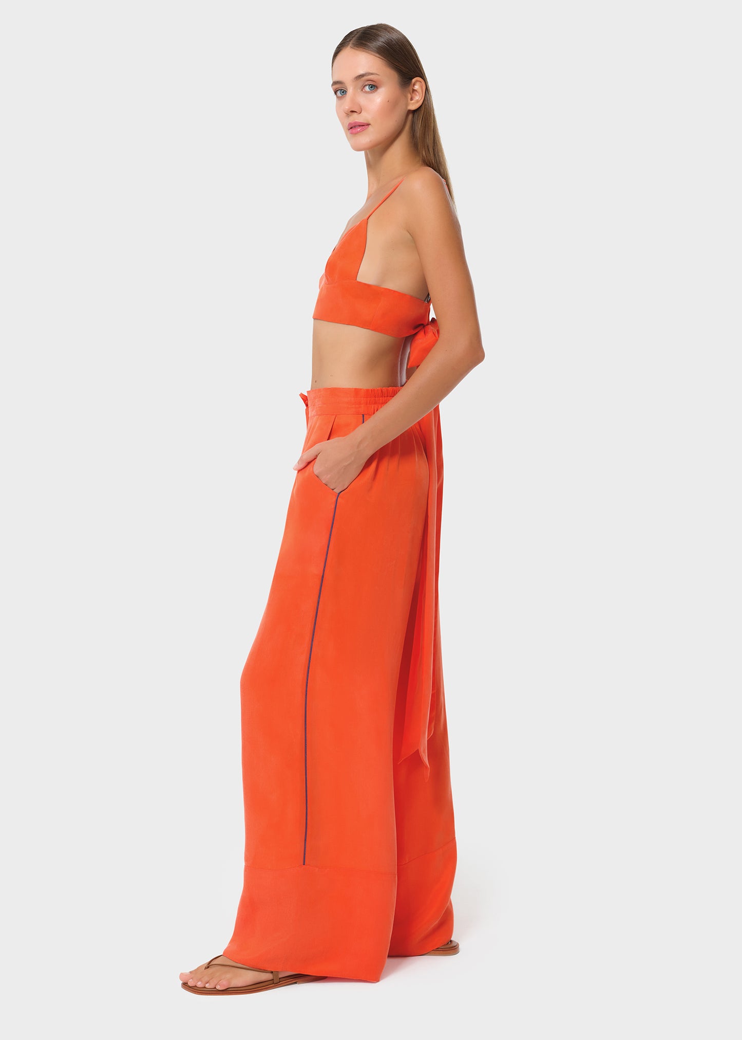 Model wearing a bright orange bralette with matching drawstring wide-leg pants, side view with hand in pocket on studio background