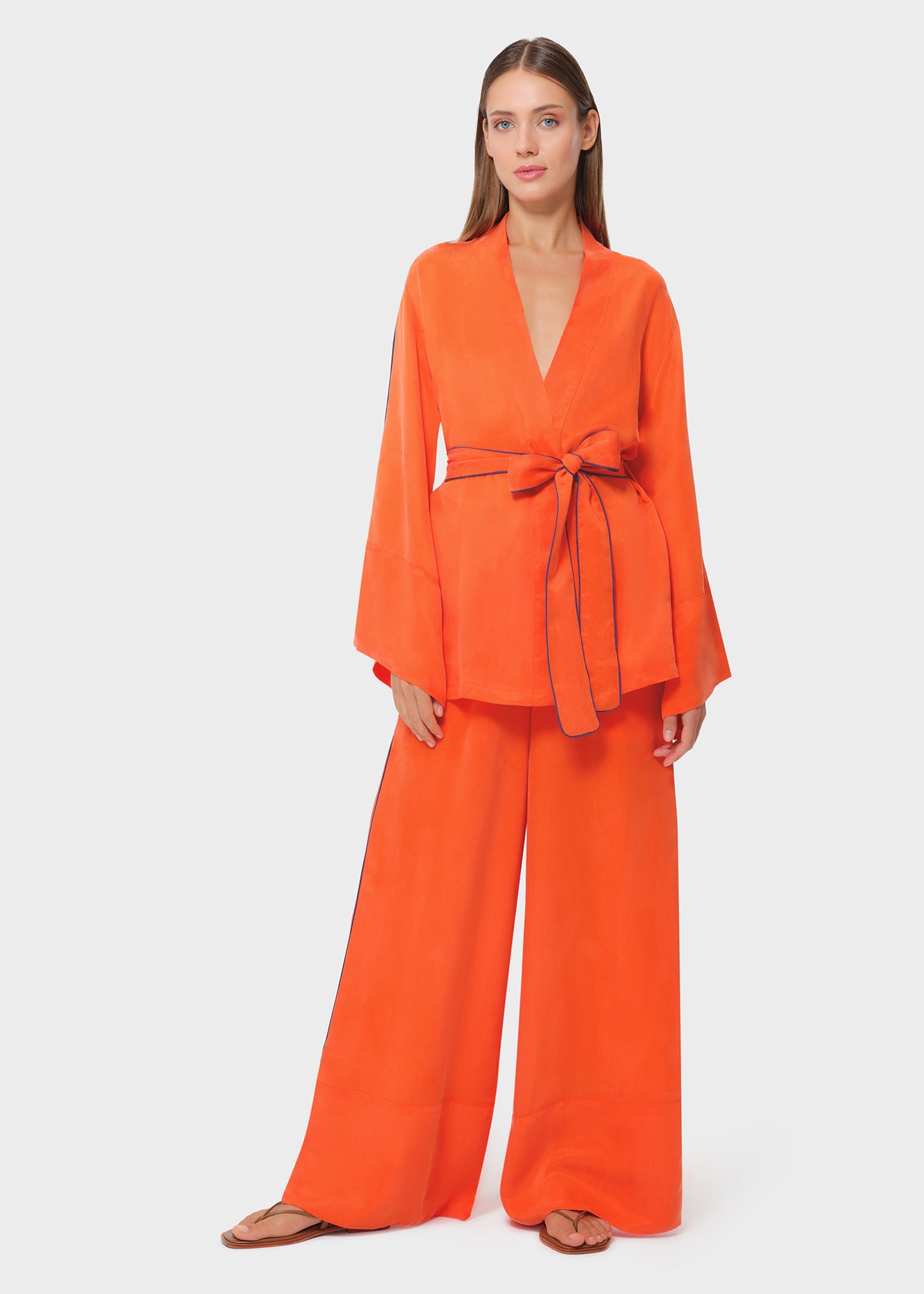 Bright orange wrap top with wide sleeves and tie belt, paired with matching wide-leg pants and sandals, studio background.