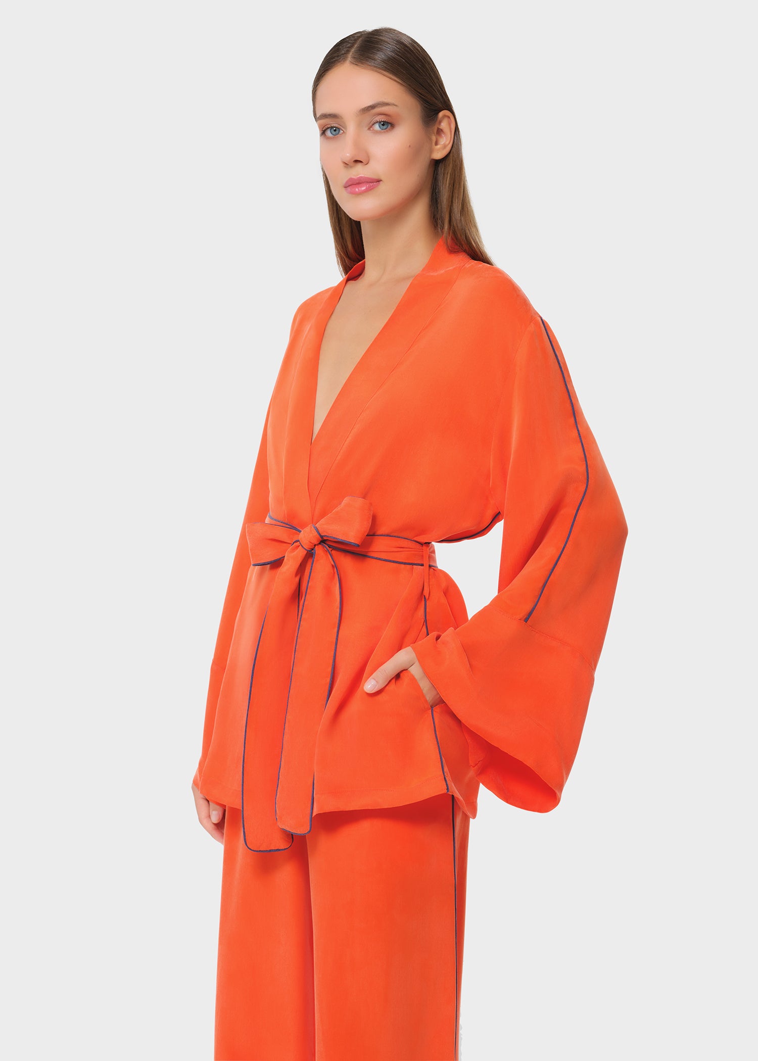 Bright orange wrap top with wide sleeves and contrast piping, tied at the waist, paired with matching wide-leg pants on a studio background.