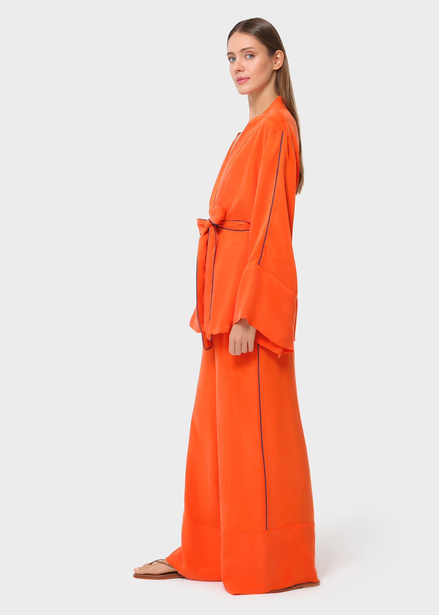 Side view of a bright orange wrap top with wide sleeves and tie belt, paired with matching wide-leg pants and flat sandals, studio background.