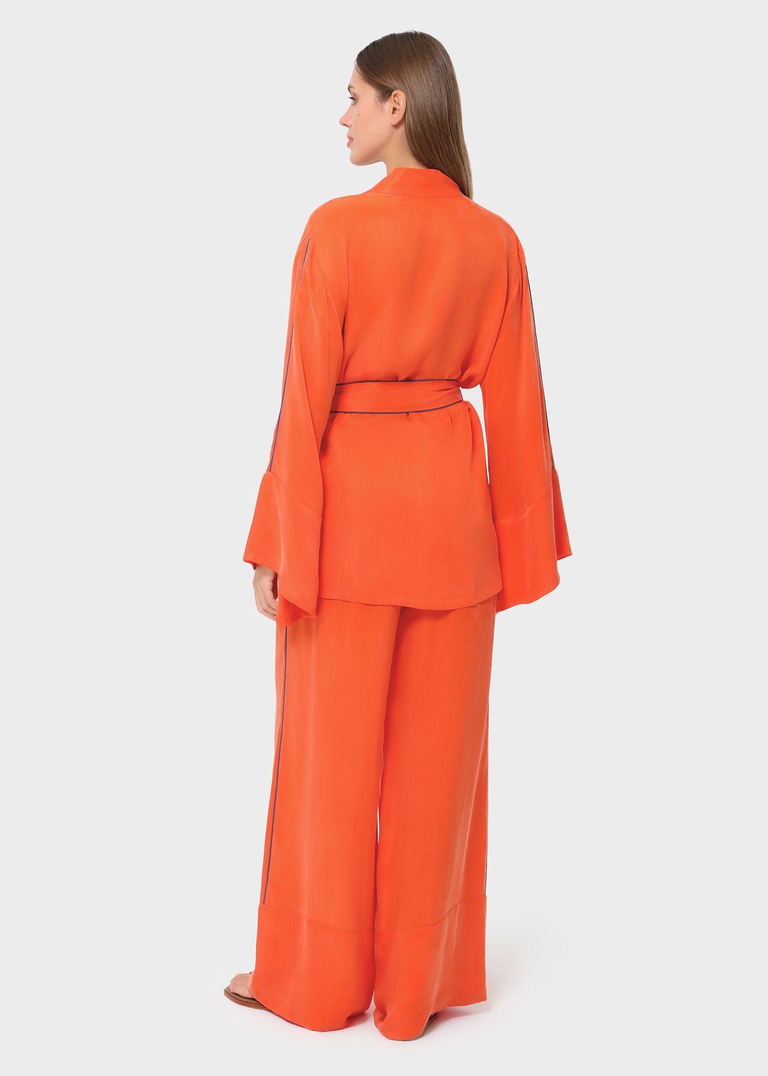 Back view of a bright orange wrap top with wide bell sleeves and matching wide-leg pants, cinched at the waist with a tie belt, styled with flat sandals on a studio background.