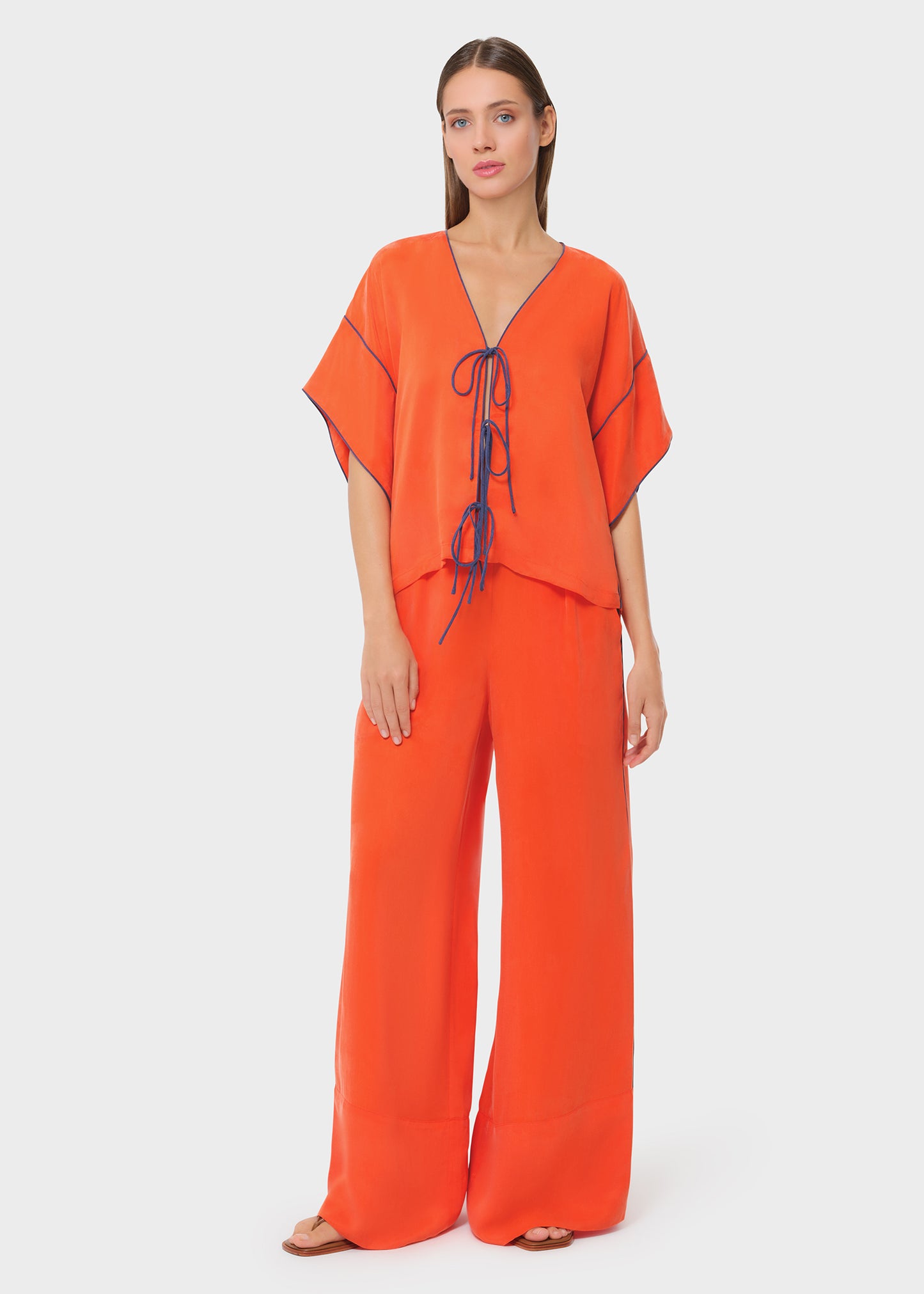 Full-length view of a bright orange co-ord set featuring a relaxed V-neck top with contrast piping and tie-up detail, paired with wide-leg flowy pants for an easy, statement look.