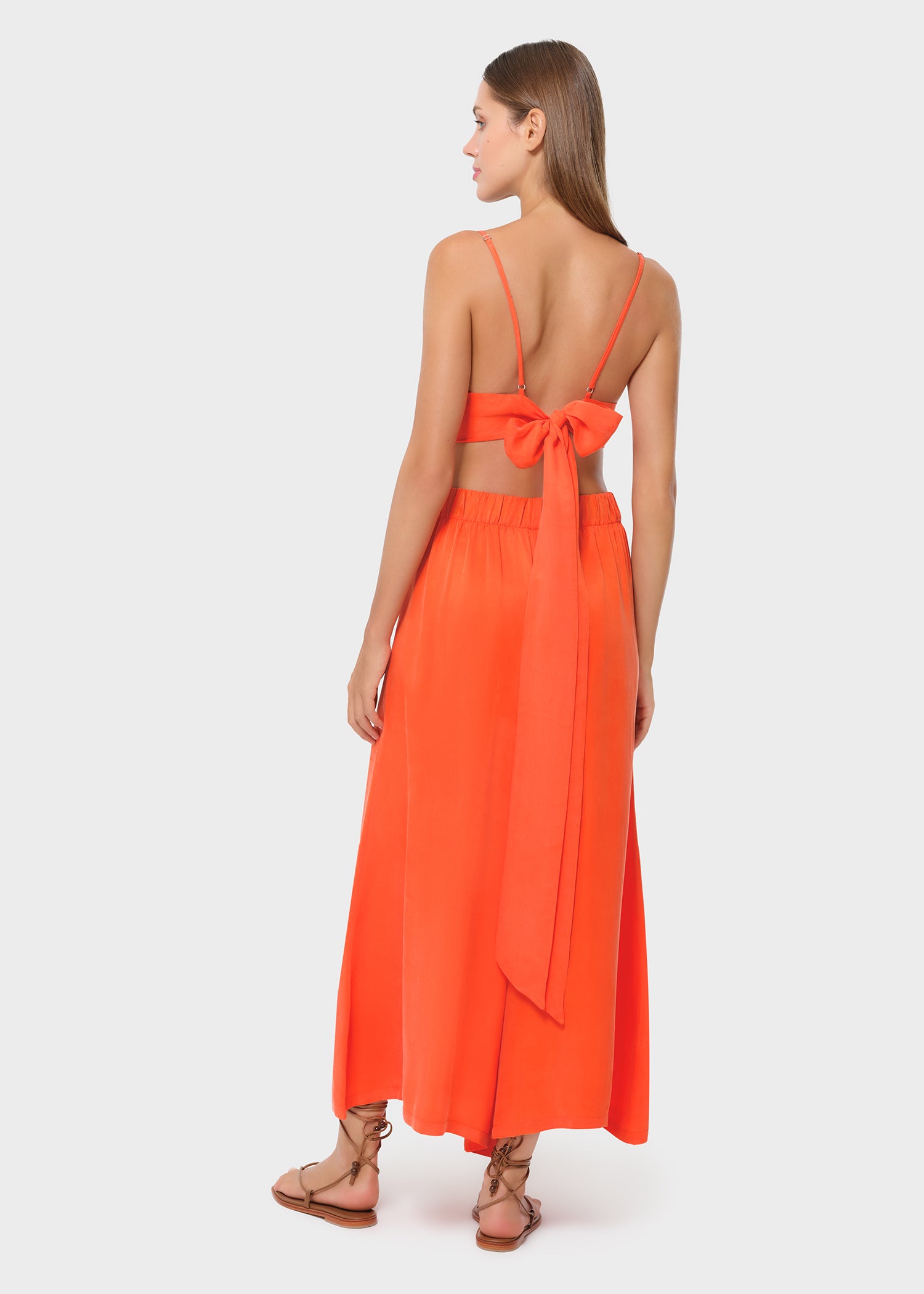 Model wearing a bright orange bralette with matching high-waisted wide-leg pants, back view with bow-tie detail on studio background