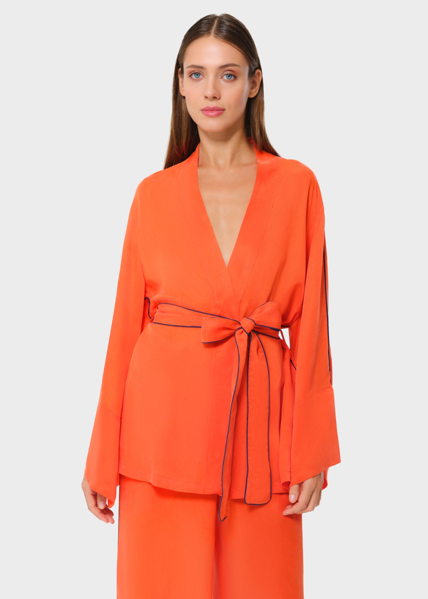 Woman wearing a vibrant orange wrap-style lounge set with wide bell sleeves and contrast navy piping, cinched at the waist with a matching tie belt, paired with coordinating wide-leg pants, studio background