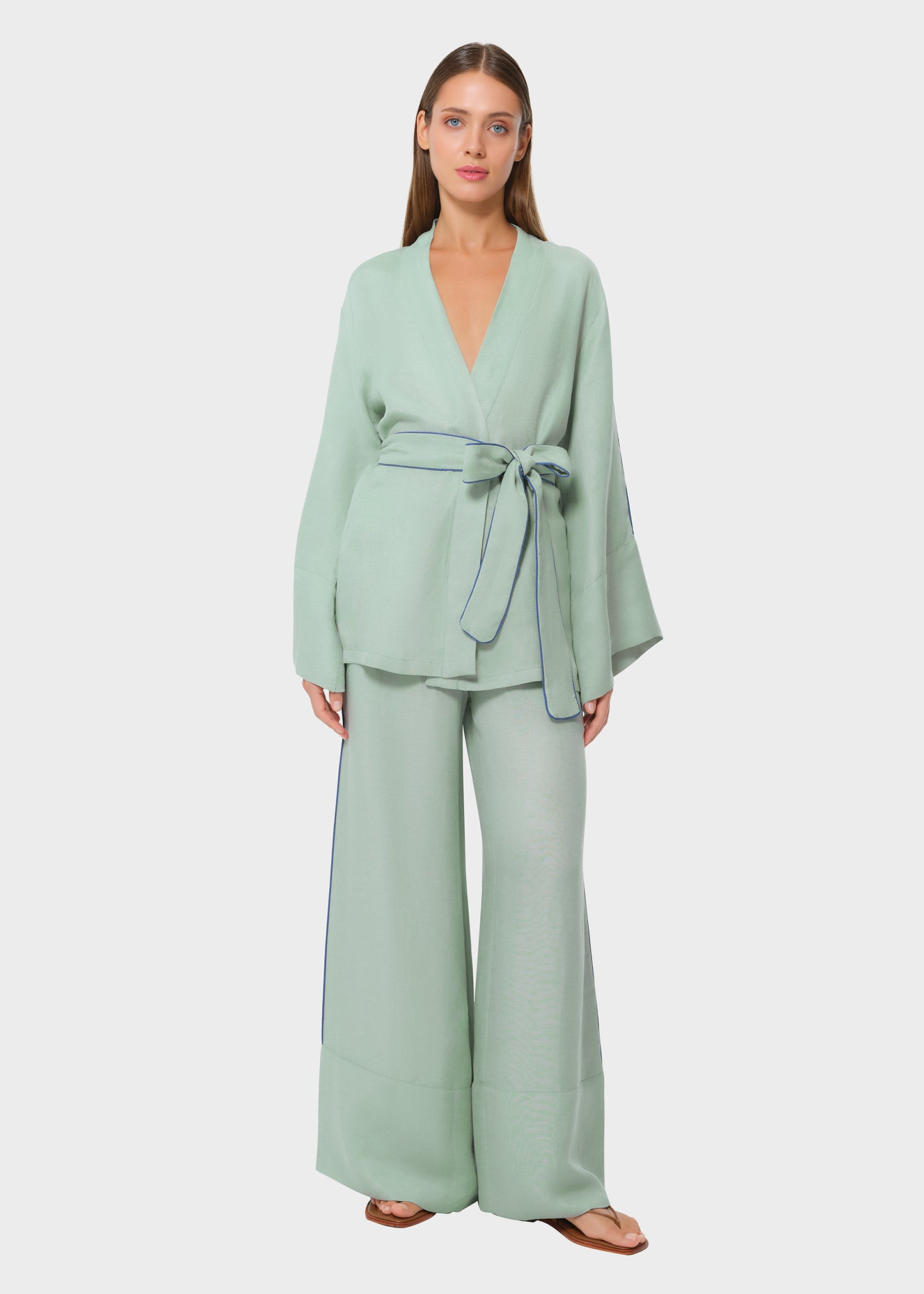 Front view of woman wearing a mint green wrap-style top with wide sleeves and blue piping detail, tied at the waist with a fabric belt, paired with matching wide-leg pants and flat sandals, studio background