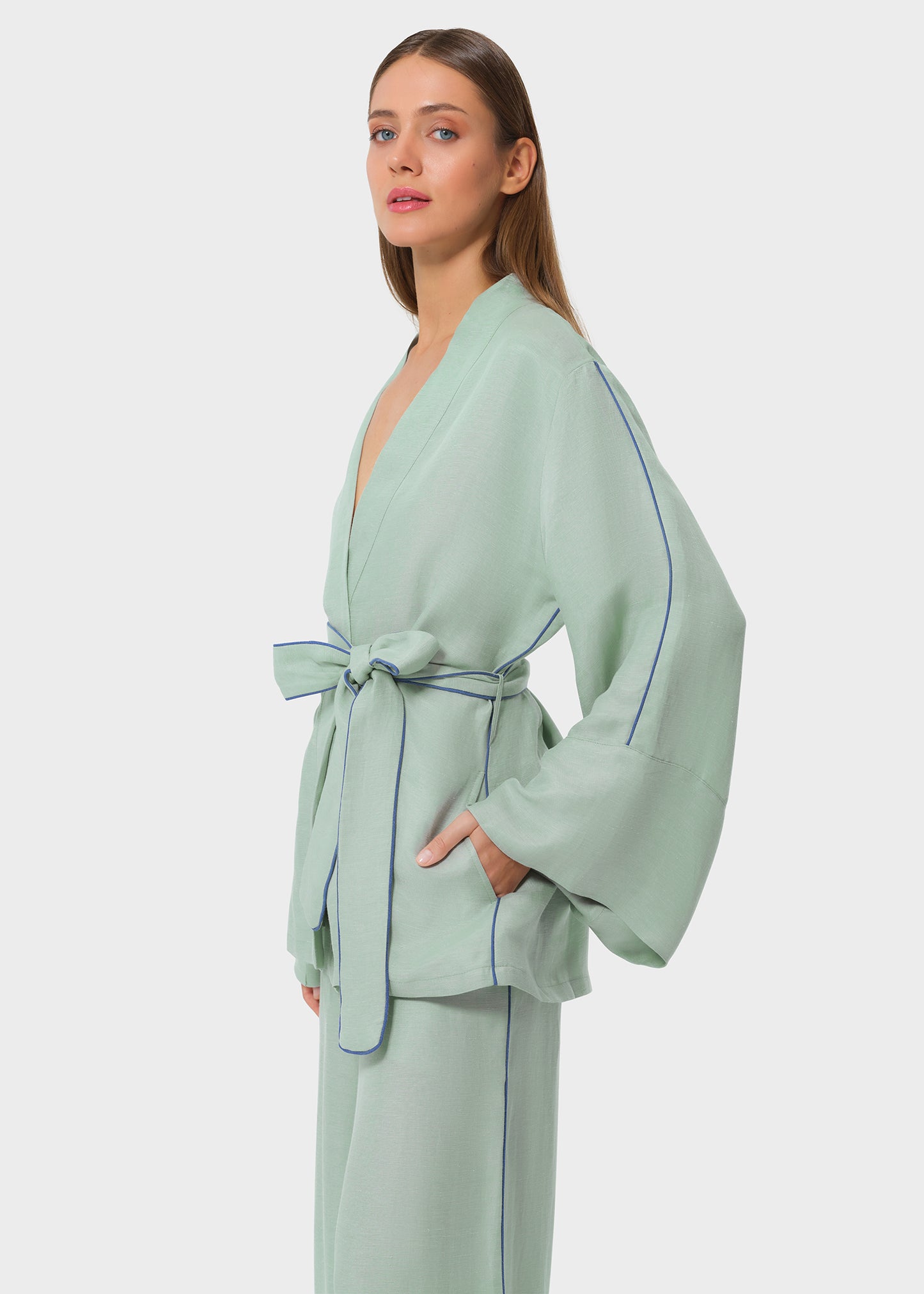 Side view of woman wearing a mint green wrap top with wide sleeves, blue piping details, and matching pants, tied at the waist with a fabric belt, studio background