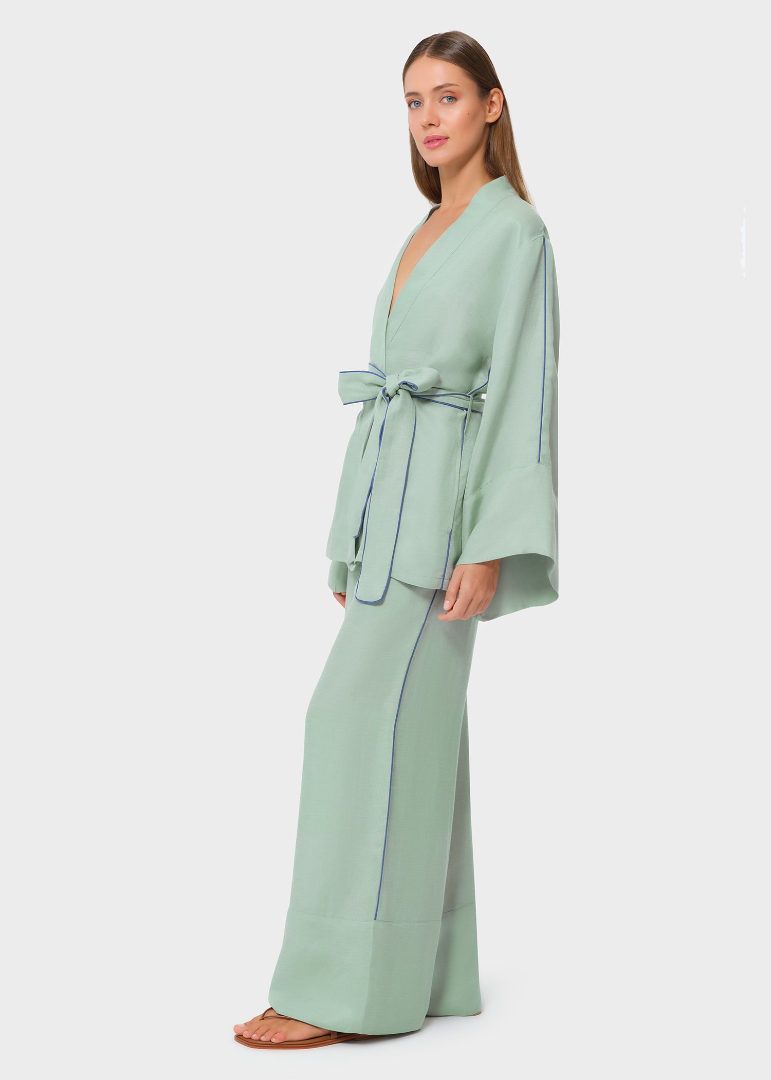 Side view of woman wearing a mint green wrap-style top with wide sleeves and blue piping detail, tied at the waist with a matching fabric belt, paired with coordinating wide-leg pants and flat sandals, studio background