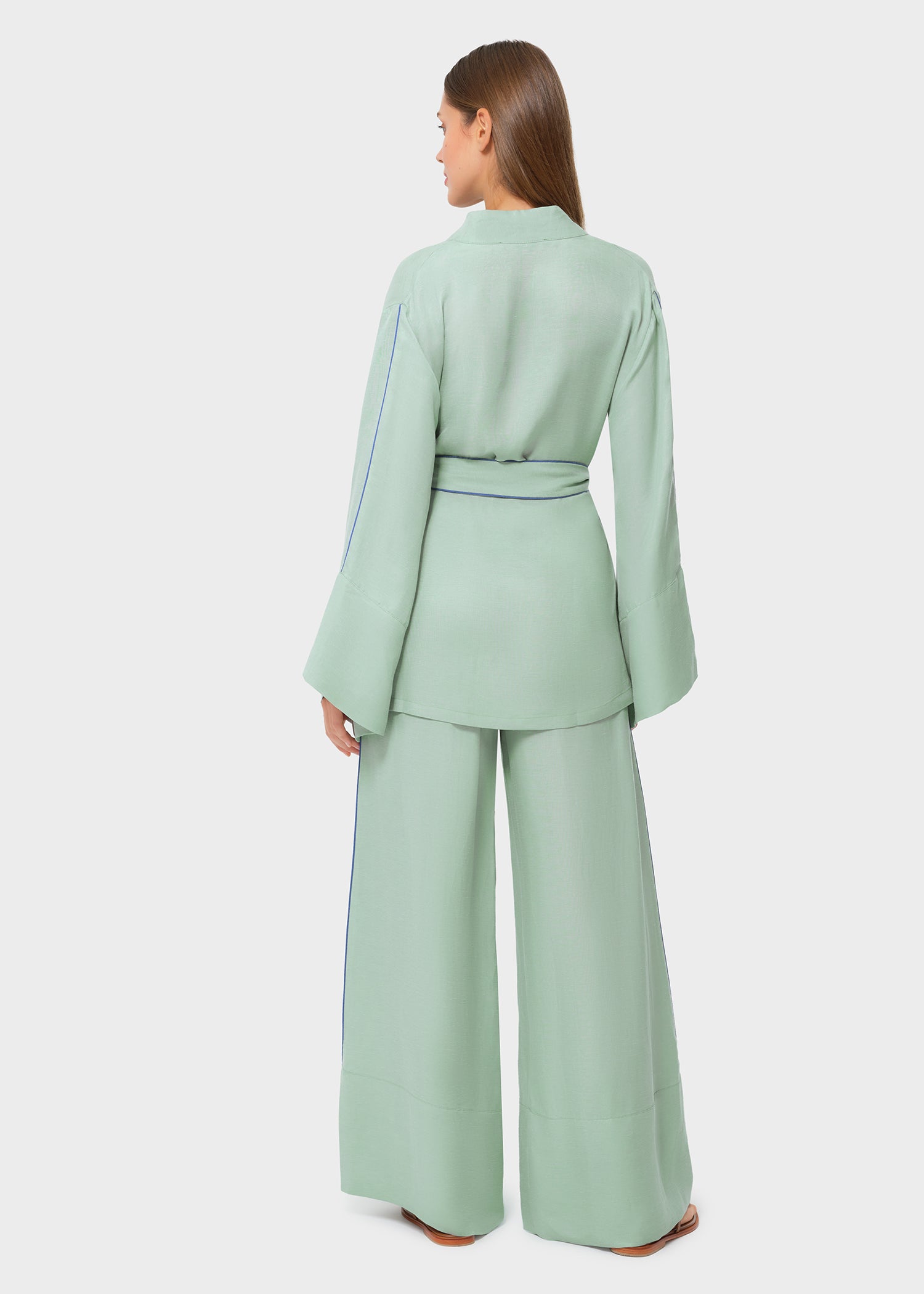 Back view of woman wearing a mint green wrap-style top with wide sleeves and blue piping detail, cinched at the waist with a matching belt, paired with coordinating wide-leg pants and flat sandals, studio background