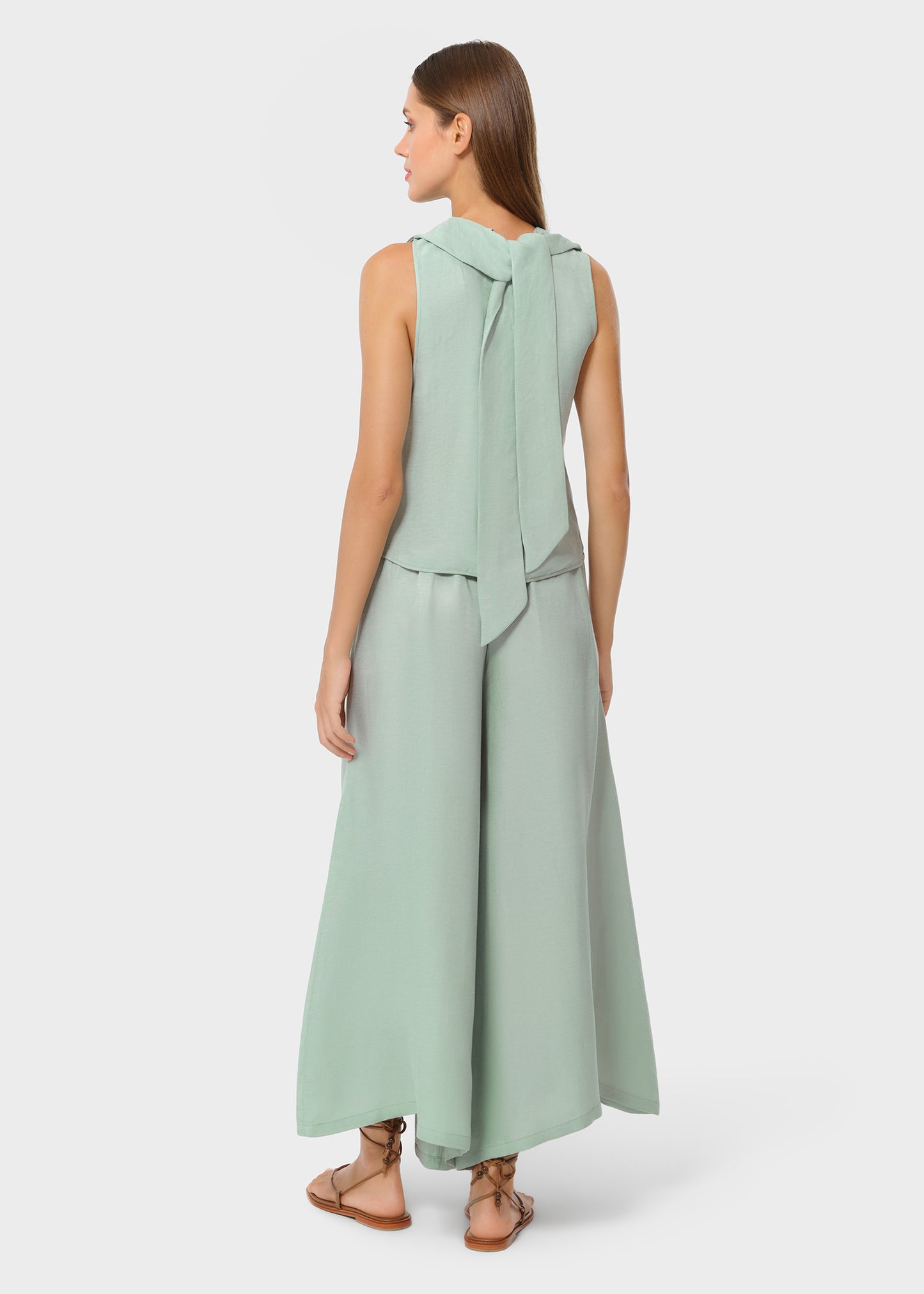 Back view of a woman in a mint sleeveless top with a tie detail at the neckline and matching wide-leg pants, styled with flat sandals in a studio setting.