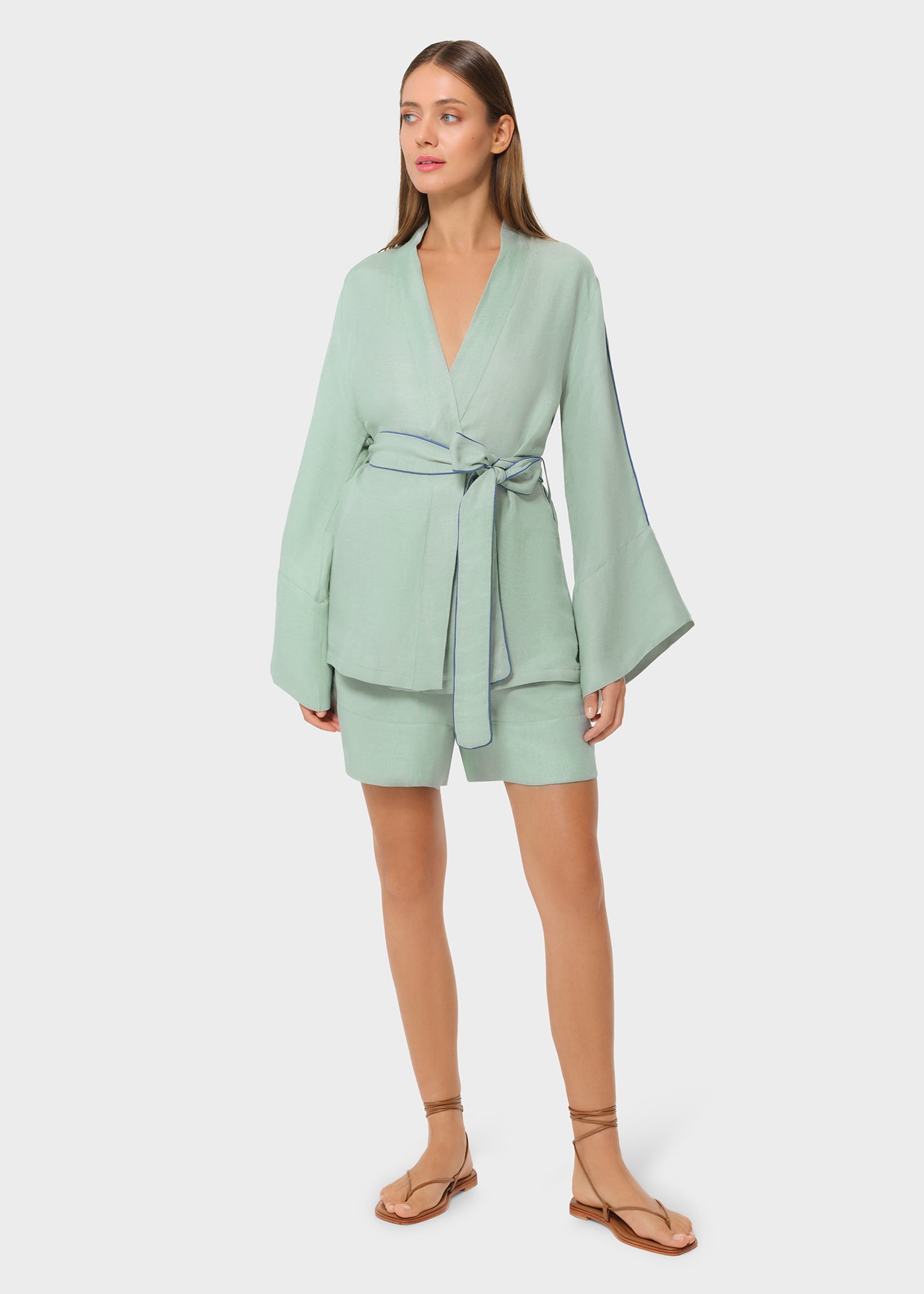 Woman wearing a mint green wrap-style top with wide bell sleeves and blue piping detail, tied at the waist with a matching belt, paired with coordinating tailored shorts and strappy flat sandals, studio background