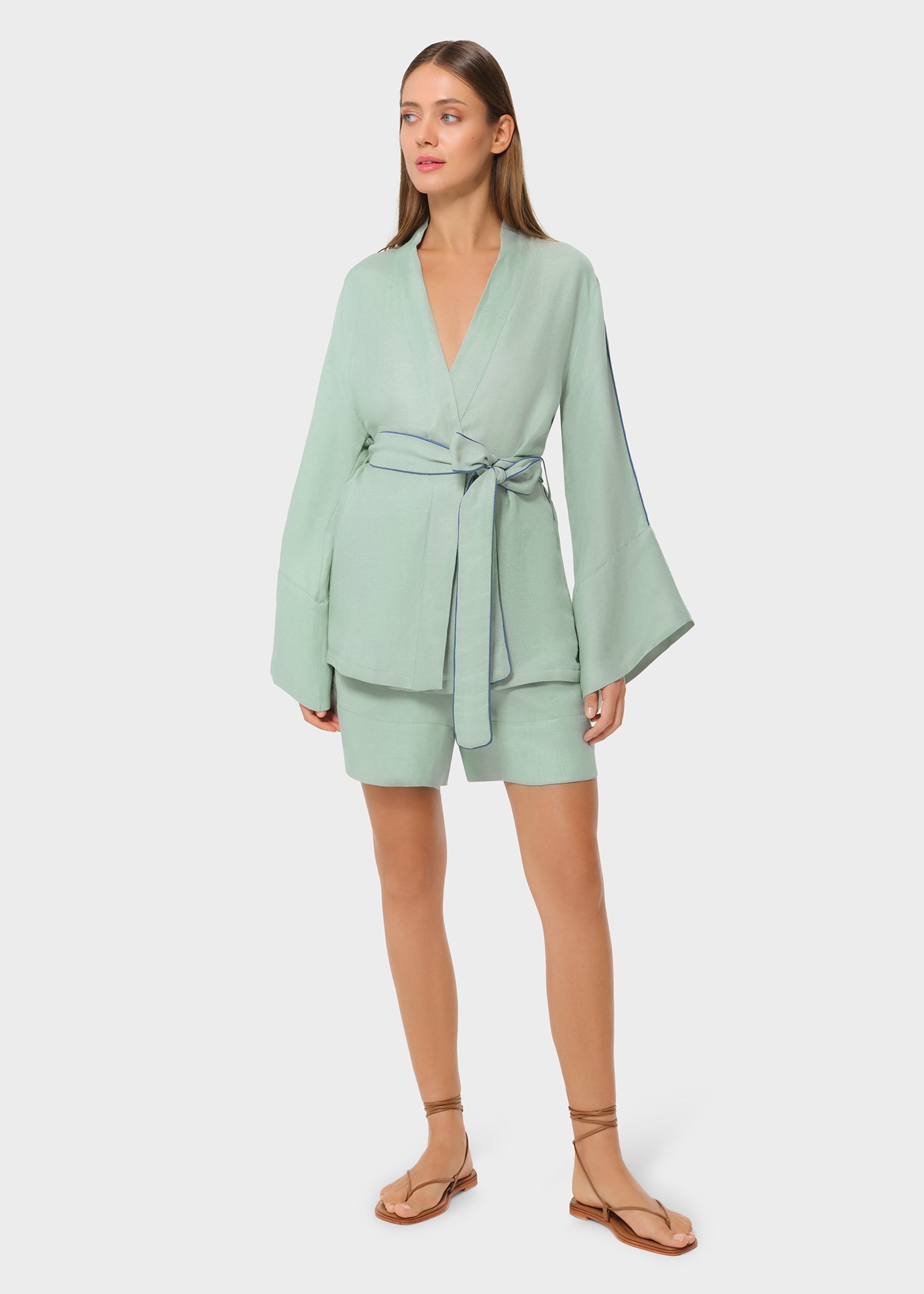 Woman wearing a mint green wrap-style top with wide bell sleeves and blue piping detail, tied at the waist with a matching belt, paired with coordinating tailored shorts and strappy flat sandals, studio background