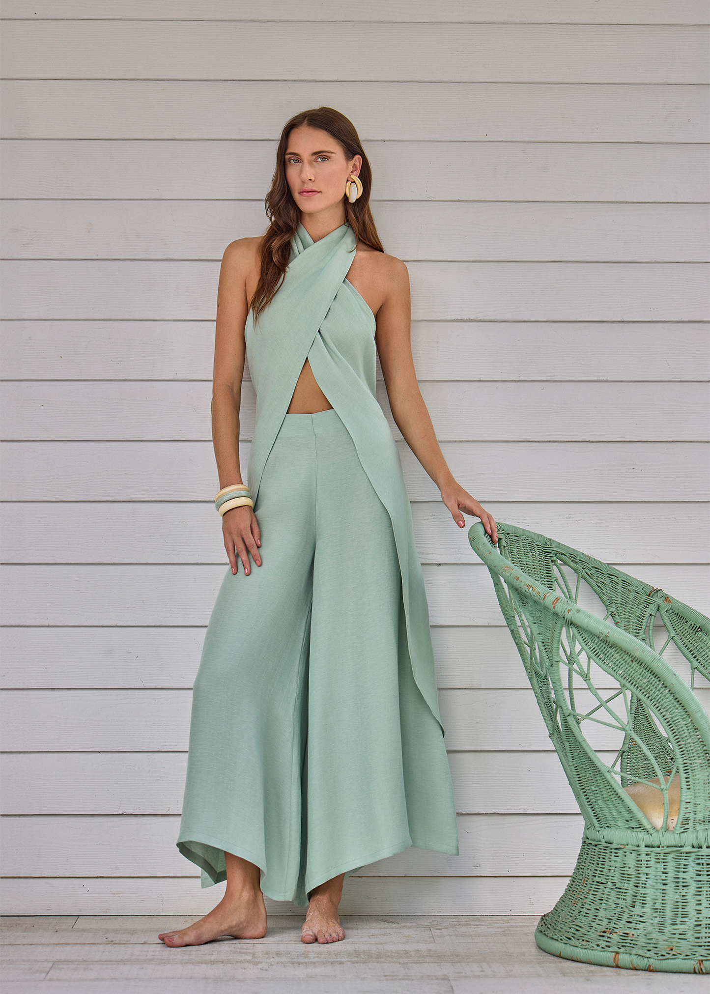 Model wearing a sage green halter wrap top with matching wide-leg pants, standing barefoot against paneled wall with woven chair nearby
