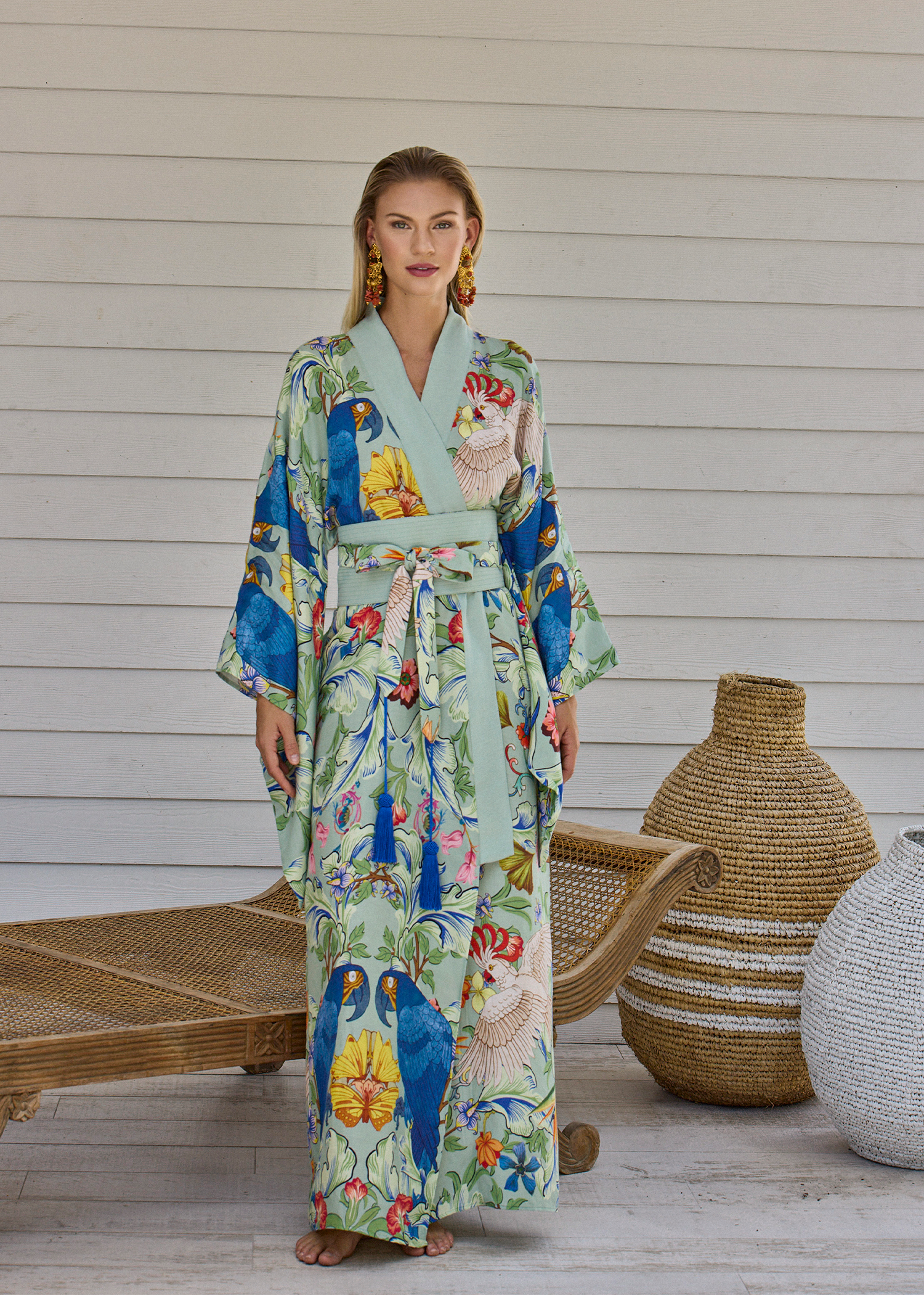 Woman wearing a green printed belted kimono maxi robe standing indoors near a lounge chair and woven floor vases.