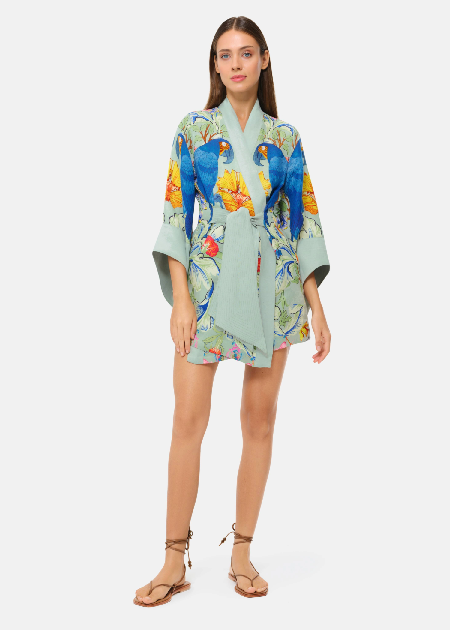Model wearing a mint green floral kimono-style mini robe with blue parrot print and tie waist.