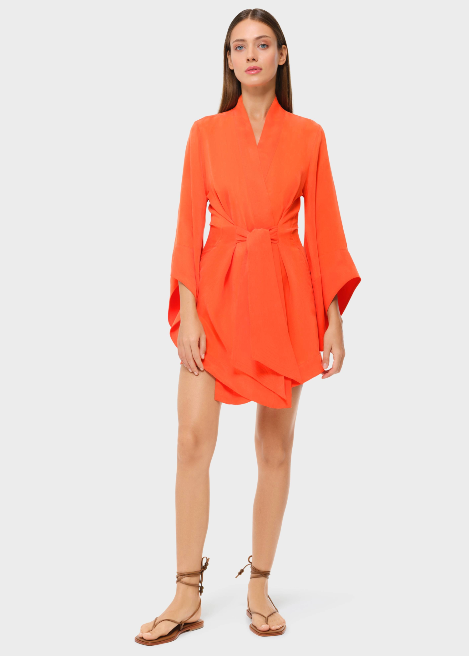 Model wearing an orange belted wrap mini dress with wide sleeves and flat sandals on a light grey background.